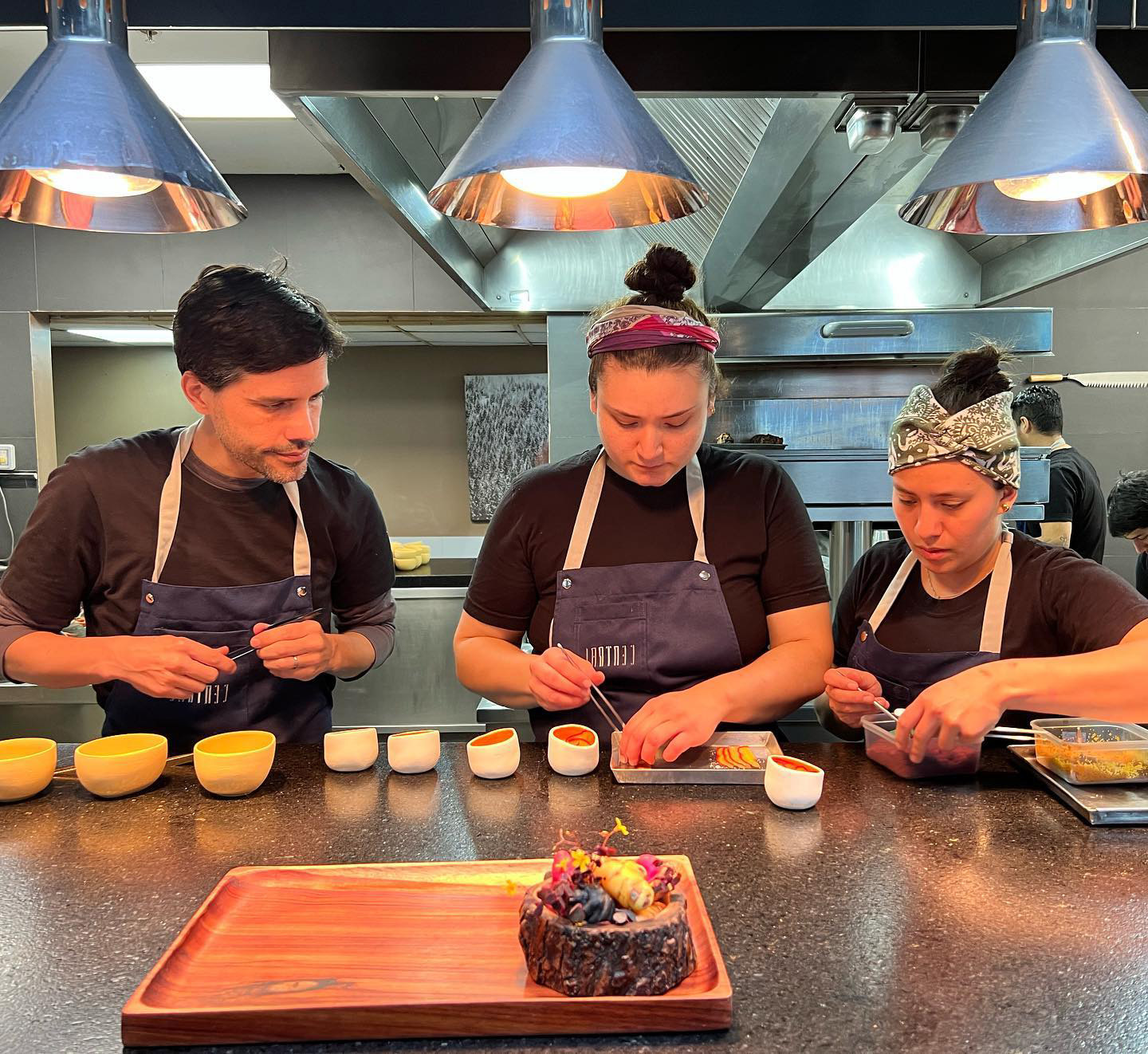 Three chefs in Lima preparing intricate dishes in a professional kitchen under bright overhead lights.