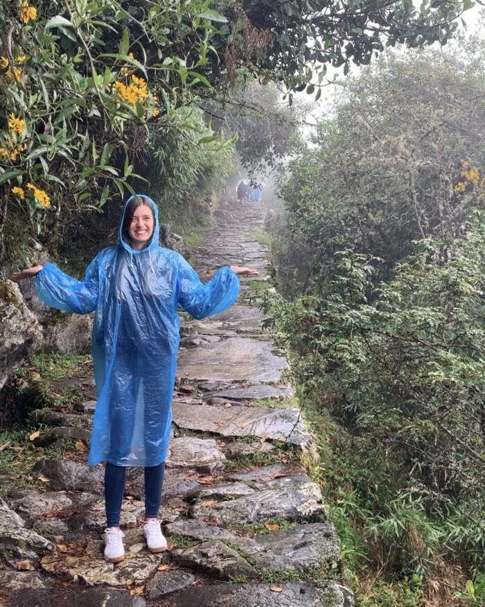 Smiling woman in a blue poncho standing on a misty stone path along the Inca Trail.