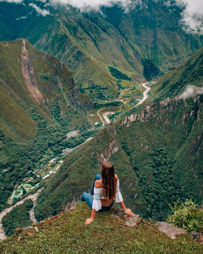 Woman sitting on a cliff edge overlooking winding rivers and green mountains.